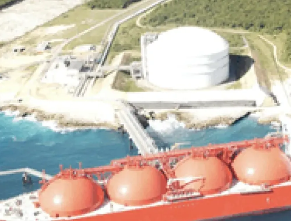 Aerial view of a large red liquefied natural gas (LNG) tanker docked at a coastal facility. The facility includes a large white storage tank and infrastructure surrounded by green vegetation. The sea is a vibrant blue, and the coastline is rocky.