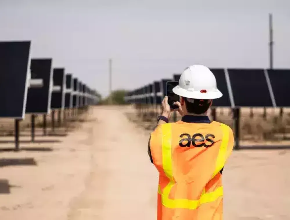 A person wearing a hard hat and an orange safety vest with 'aes' on the back stands in a solar panel field, taking a photo with a smartphone. Rows of solar panels extend into the distance on both sides.