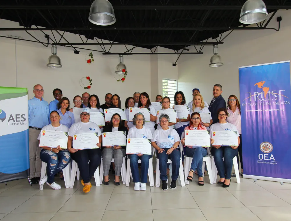 A group of people posing with certificates in a room with banners from AES Puerto Rico and The Trust for the Americas.