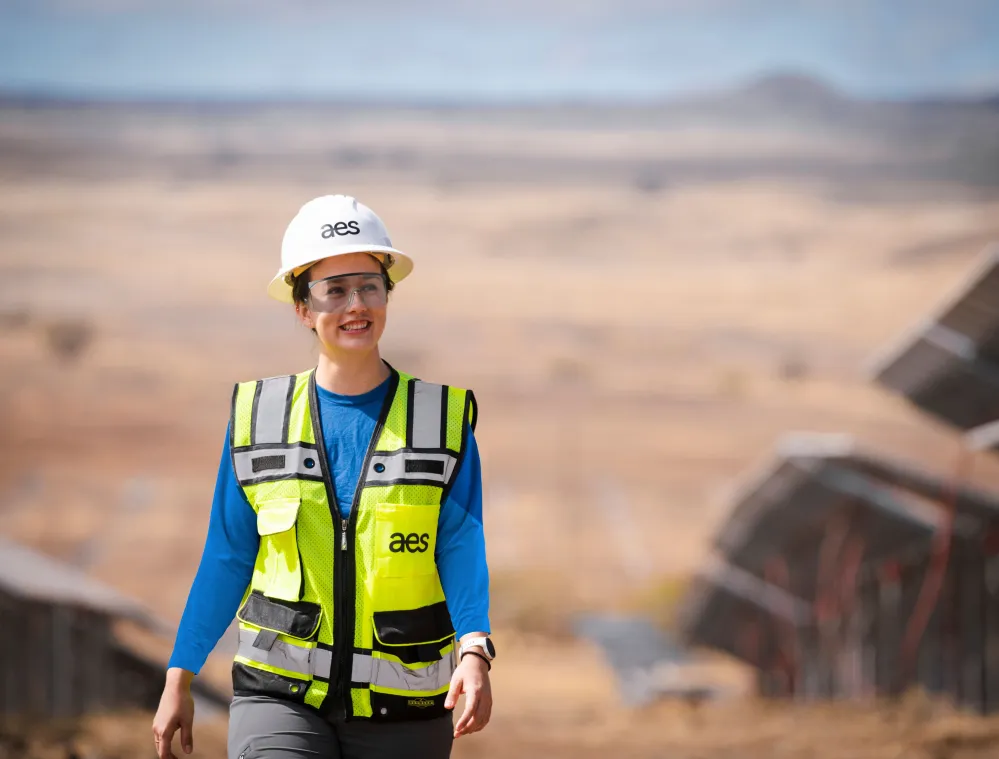 A person wearing a white hard hat and a high-visibility vest with 'aes' logo walks outdoors near solar panels in a desert landscape.