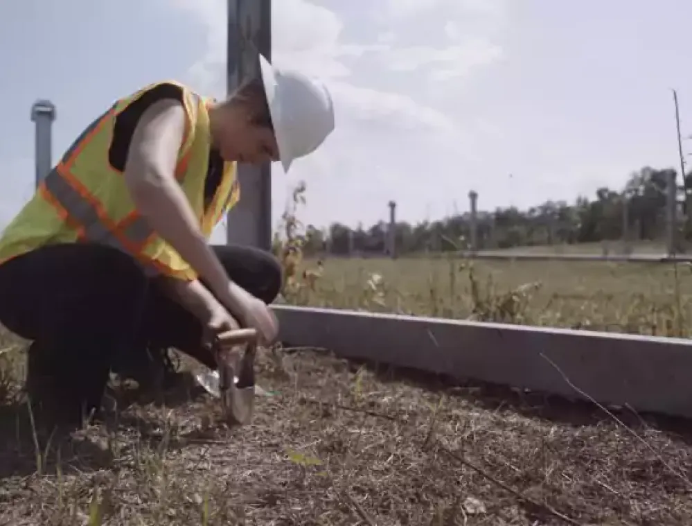 A person wearing a white hard hat and orange safety vest crouches down, using a small tool to dig into the ground near a concrete border in a grassy area. Several tall poles are visible in the background under a partly cloudy sky.