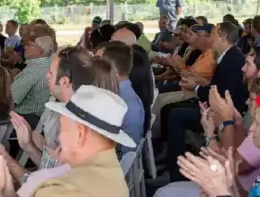 A large group of people seated under a tent, clapping their hands. The audience is diverse, with individuals of various ages and attire, including hats and casual clothing. The setting appears to be an outdoor event with trees visible in the background.
