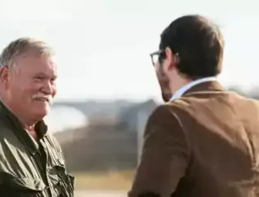 Two men are standing outdoors, engaged in conversation. One man, wearing a dark green jacket, is smiling. The other man, seen from behind, is wearing a brown jacket and glasses. The background is blurred, showing an outdoor setting with some structures.