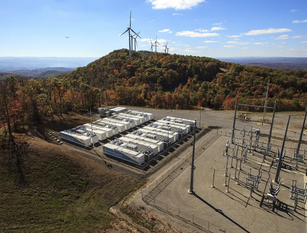 Aerial view of a wind farm on a hill with several wind turbines surrounded by autumn-colored trees. In the foreground, there is an array of white container units and electrical infrastructure on a cleared area. The landscape extends into distant hills under a clear blue sky.