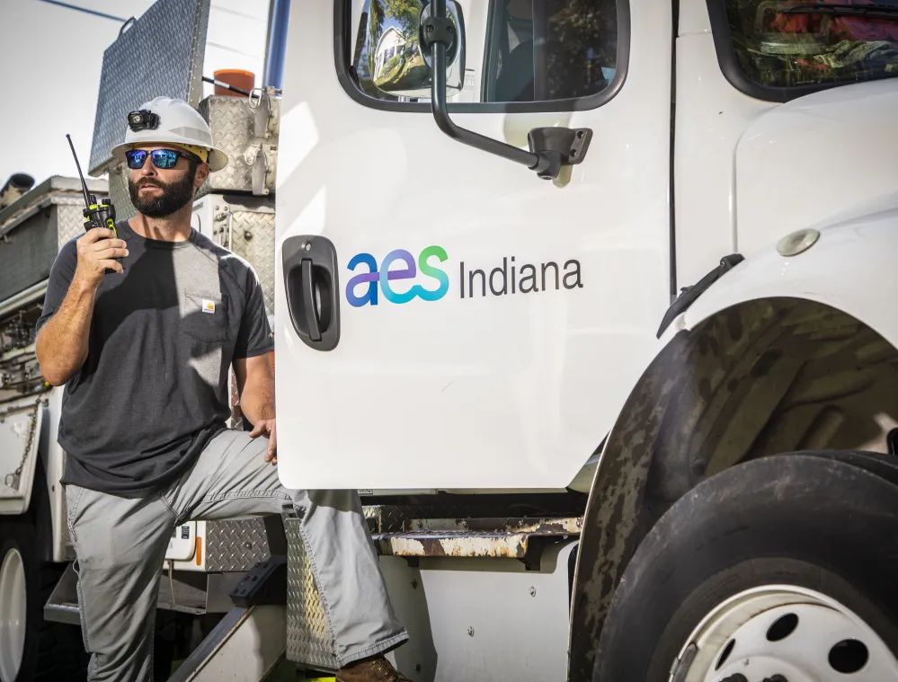 A worker wearing a hard hat and sunglasses holds a radio while leaning on a utility truck with 'aes Indiana' logo.