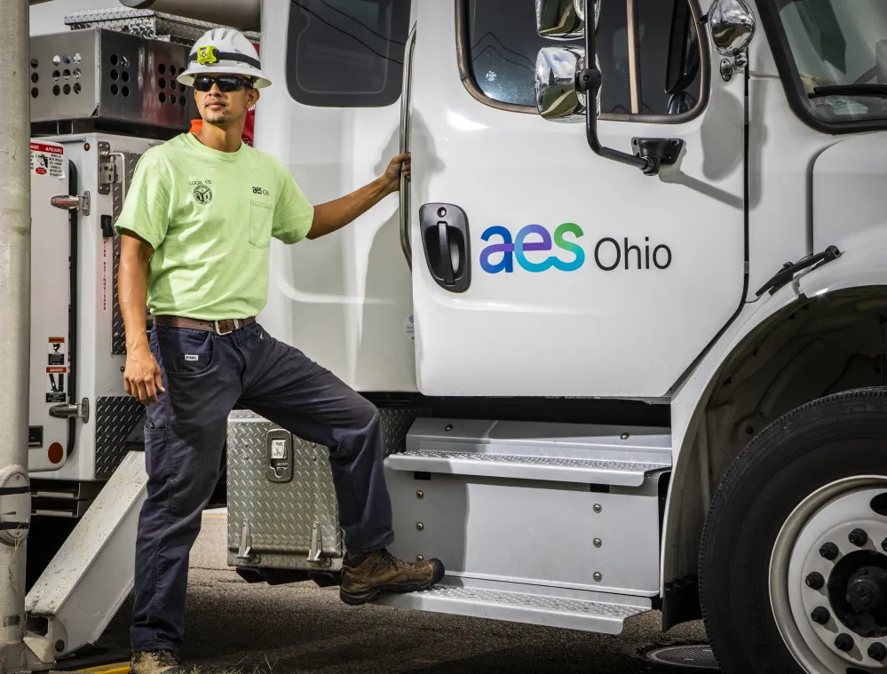 A worker wearing a hard hat and safety glasses stands on the step of a white truck with the AES Ohio logo. The worker is wearing a neon green shirt and dark pants.