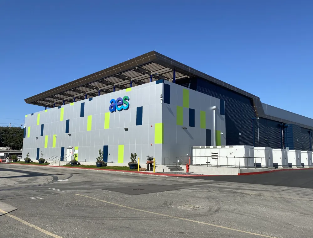 A modern building with a facade featuring a pattern of blue and green panels, displaying the 'aes' logo. The building is surrounded by a paved area and has several white utility boxes along its side. The sky is clear and blue.