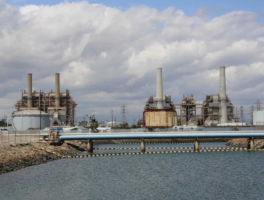 Industrial facility with several large chimneys and structures, situated by a body of water. A blue pipeline runs across the foreground. The sky is cloudy.