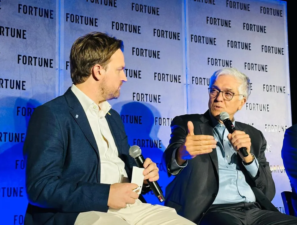 Two men seated on stage engaged in conversation, holding microphones. The background features a wall with the word 'FORTUNE' repeated. The man on the right gestures with his hand while speaking.