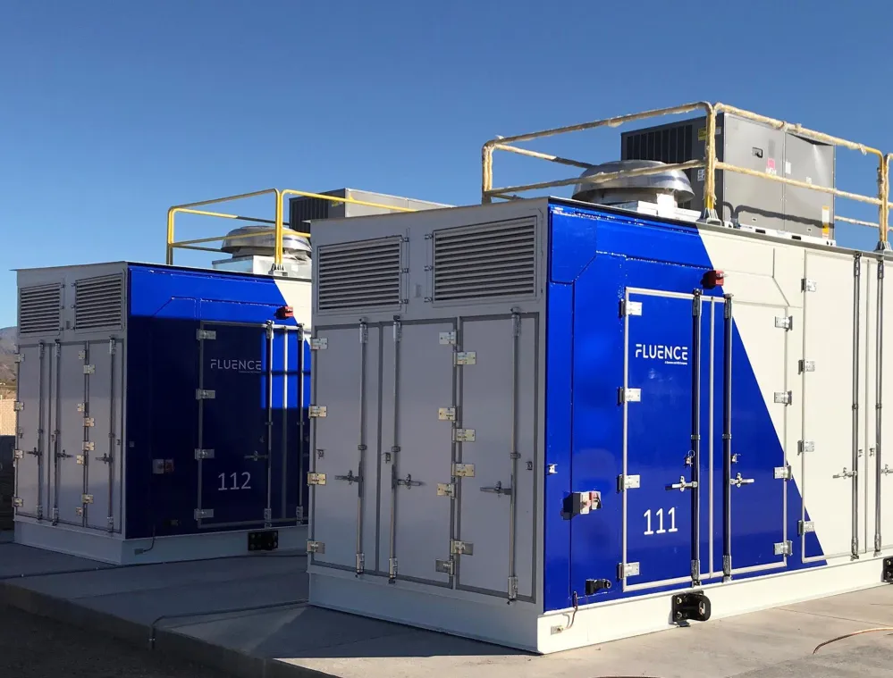 Two large industrial battery storage units with blue and white exteriors, labeled 'Fluence' and numbered 111 and 112, are situated outdoors on a concrete platform under a clear blue sky.