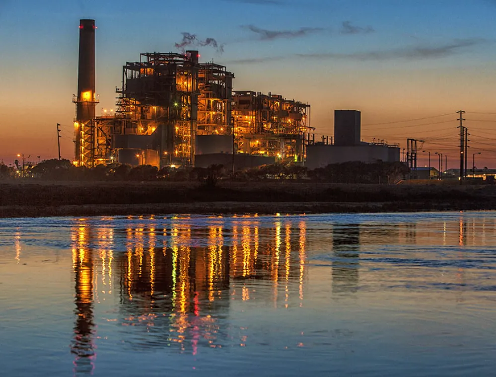 A power plant illuminated at dusk, with bright lights contrasting against the darkening sky. The plant's lights reflect in the calm water in the foreground, creating a colorful mirrored effect. The sky transitions from orange near the horizon to deep blue at the top.
