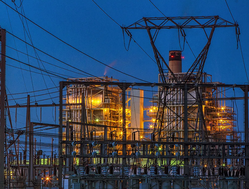 An industrial power plant is illuminated at dusk, with bright lights highlighting its structures and smokestack. The foreground is filled with a network of power lines and metal frameworks against a deep blue sky.