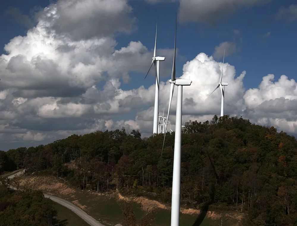 Wind turbines on a hilltop surrounded by trees, under a partly cloudy sky.