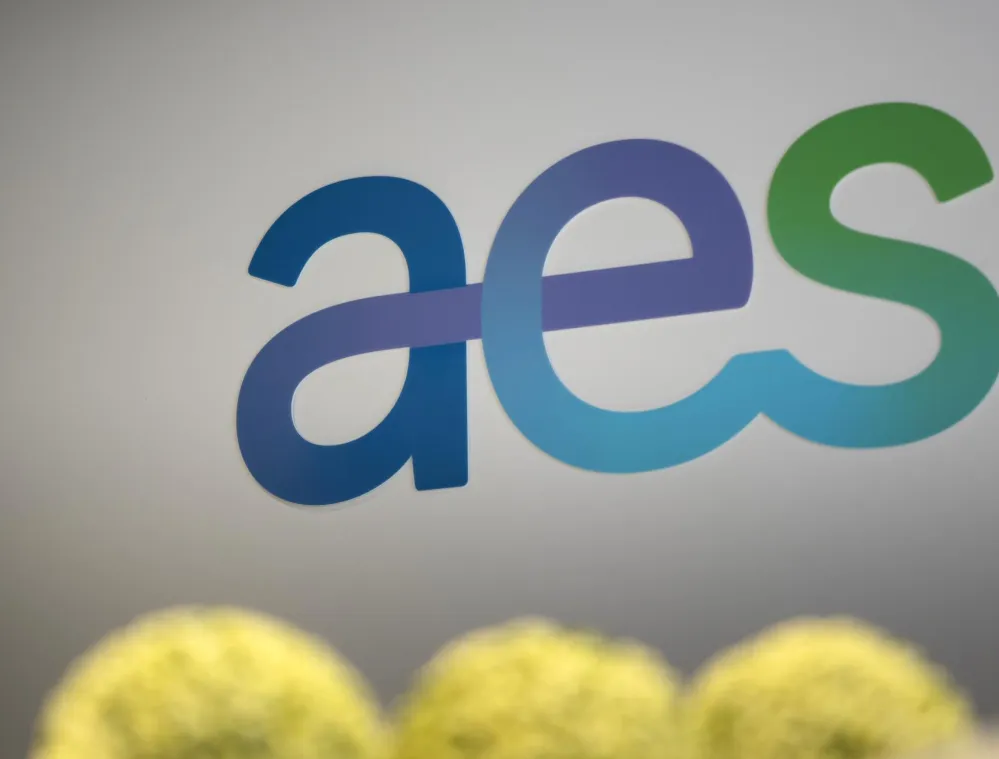 The image shows the AES logo in gradient colors of blue, purple, and green on a light gray background. The letters 'a', 'e', and 's' are connected in a smooth, overlapping design. In the foreground, there are blurred yellow-green shapes, possibly plants.