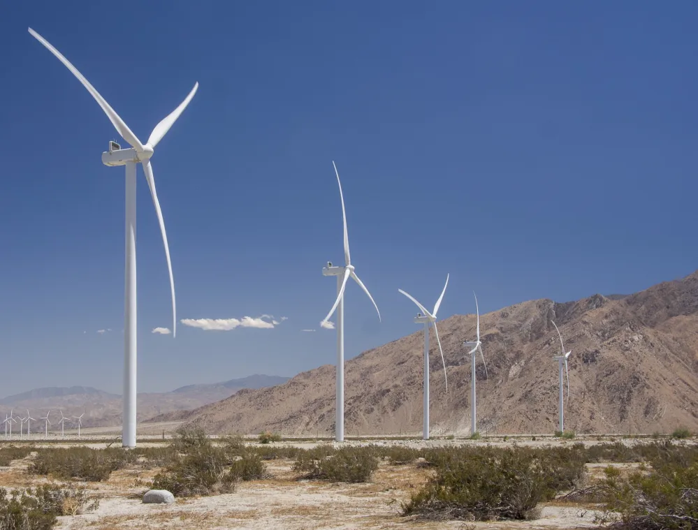 A row of white wind turbines stands in a desert landscape with sparse vegetation. The turbines are set against a backdrop of brown, rocky mountains under a clear blue sky.