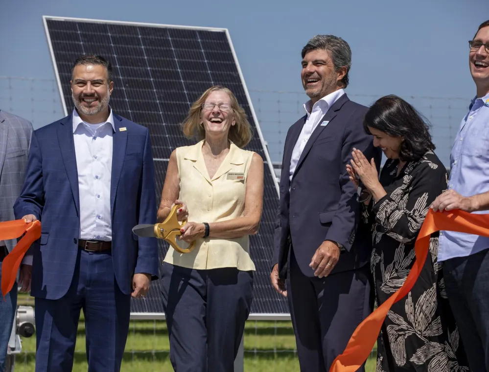 A group of people smiling and holding an orange ribbon at a ribbon-cutting ceremony in front of a solar panel. A woman in a yellow blouse is holding large scissors. The sky is clear and blue.