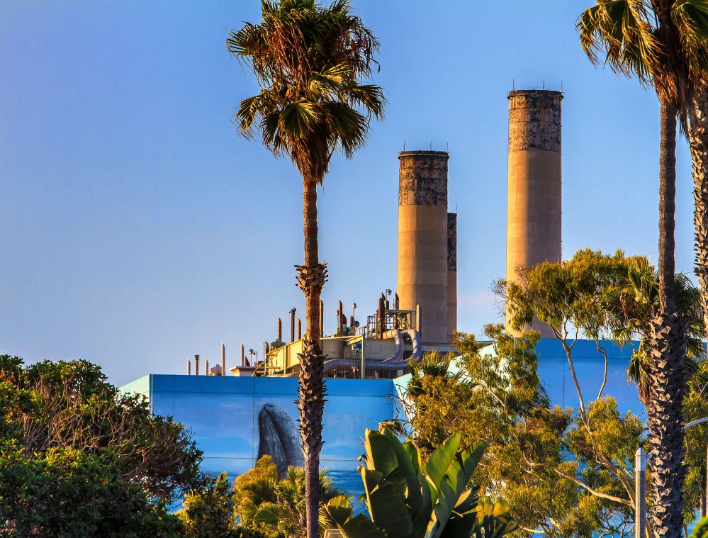 A power plant with two tall smokestacks is partially obscured by palm trees and other vegetation. The structure has a blue facade with industrial pipes visible on top. The sky is clear and blue, suggesting a sunny day.