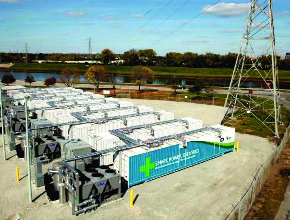 Aerial view of a power facility with multiple white containers and electrical equipment. A large transmission tower stands nearby, with a road curving around the site. Trees and a river are visible in the background under a partly cloudy sky.