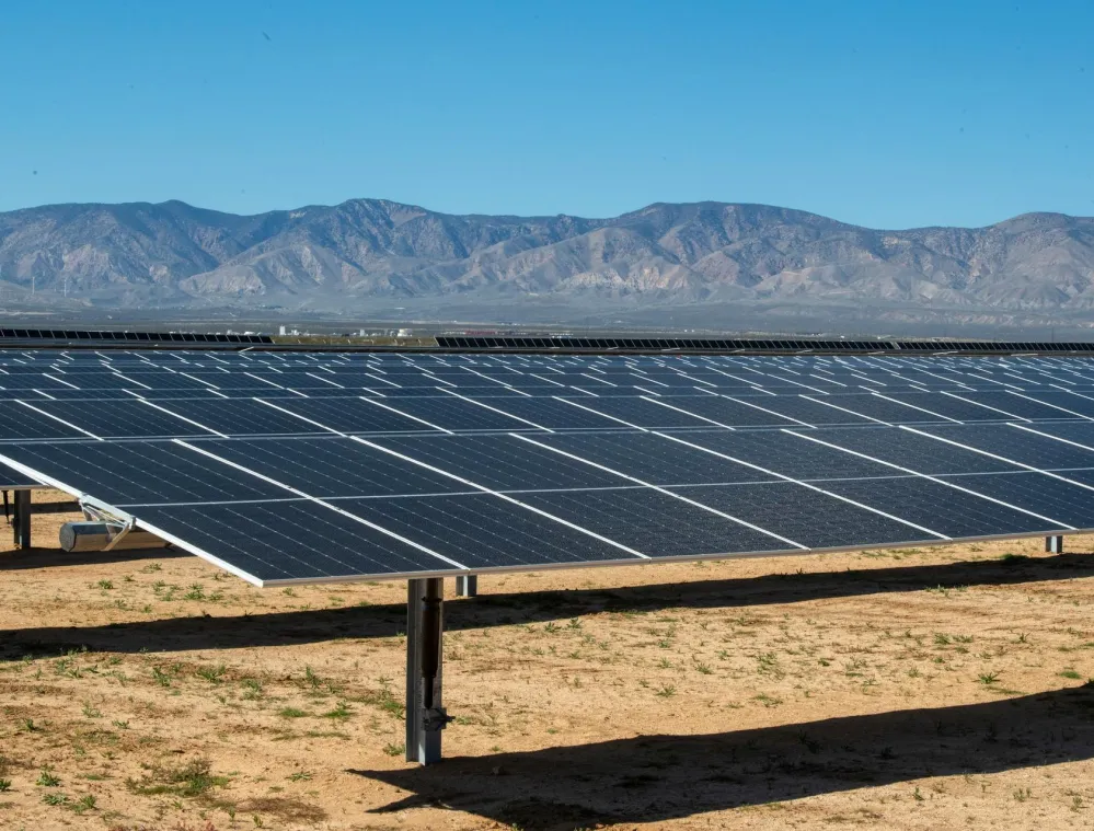 A large array of solar panels is installed on a flat, dry landscape under a clear blue sky. In the background, a range of mountains is visible, adding depth to the scene.