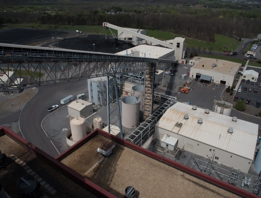 Aerial view of an industrial facility with large storage tanks, a conveyor system, and several buildings. The surrounding area includes parking lots and a green landscape with trees in the distance.