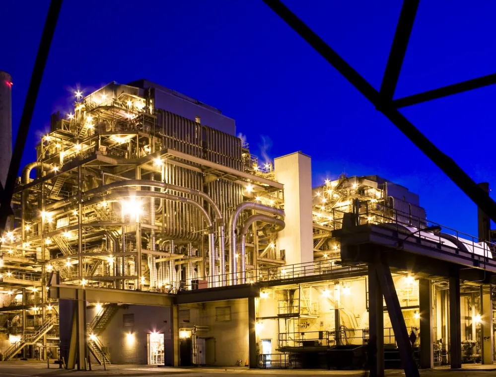 Illuminated industrial plant with complex piping and structures, set against a deep blue evening sky. The plant is brightly lit with numerous lights, highlighting the intricate network of pipes and platforms. A tall chimney is visible in the background.