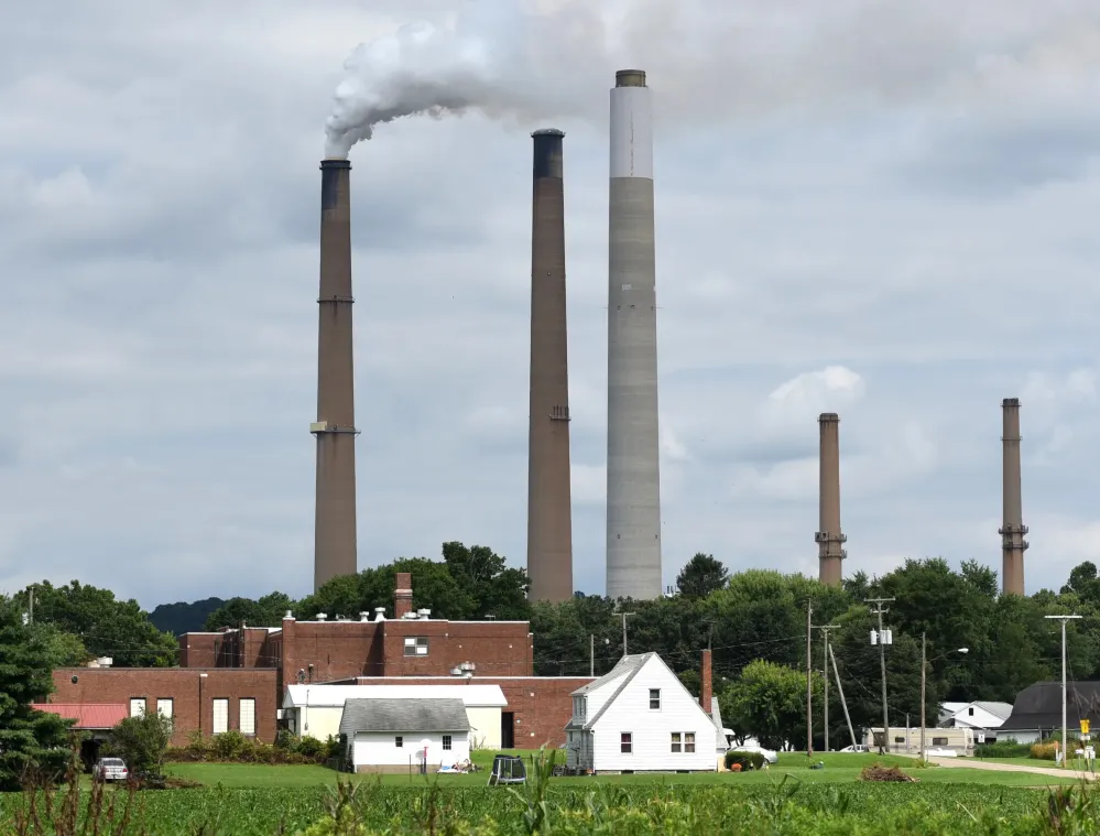 A rural landscape with several tall smokestacks emitting smoke against a cloudy sky. In the foreground, there are small buildings and houses surrounded by greenery and trees.