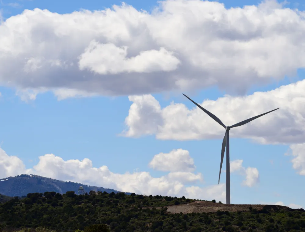 A wind turbine stands on a hilltop under a blue sky with scattered white clouds. The landscape includes a distant mountain range and green vegetation on the hills.
