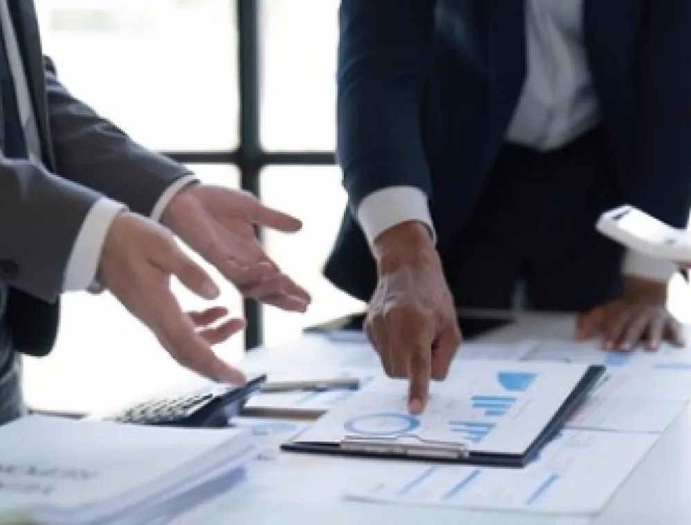 Business professionals stand around a table with documents and a tablet, discussing data shown in charts and graphs. One person points to a document with a pen, while another holds a smartphone.