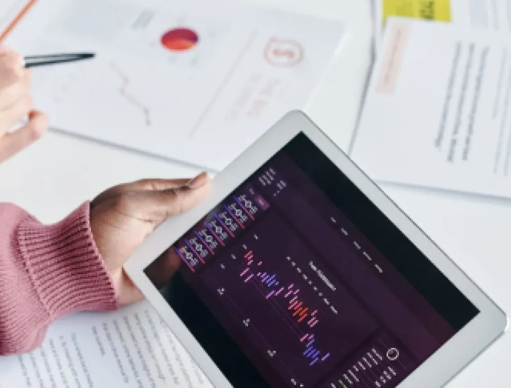 Two people reviewing financial data. One holds a tablet displaying a colorful bar chart and line graph. The other writes on a document with charts and graphs on a table.