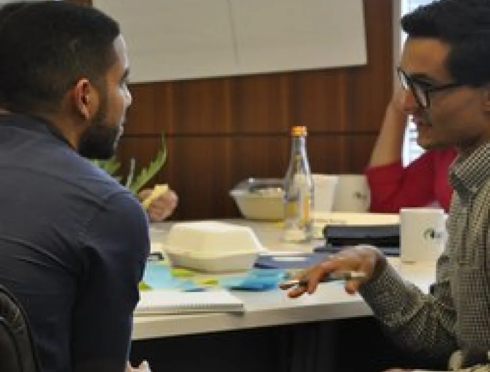 Two men sitting and having a discussion at a meeting table with notebooks, cups, and a bottle, while another person listens in the background.