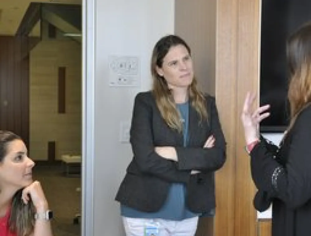 Three women are having a discussion in a modern office. One woman is seated at a table, while two others stand nearby, one gesturing with her hands. They appear engaged in conversation, with office furniture and a hallway visible in the background.