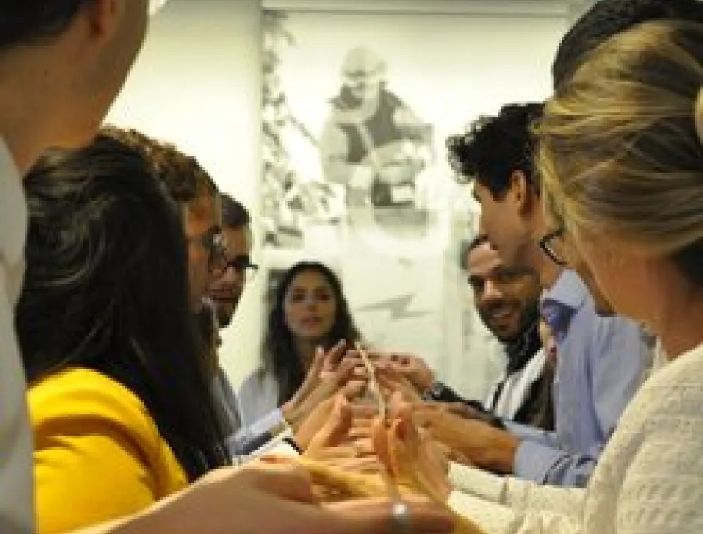 A group of people standing in two lines facing each other, participating in a team-building activity indoors, focusing on balancing a thin rod with their fingers.