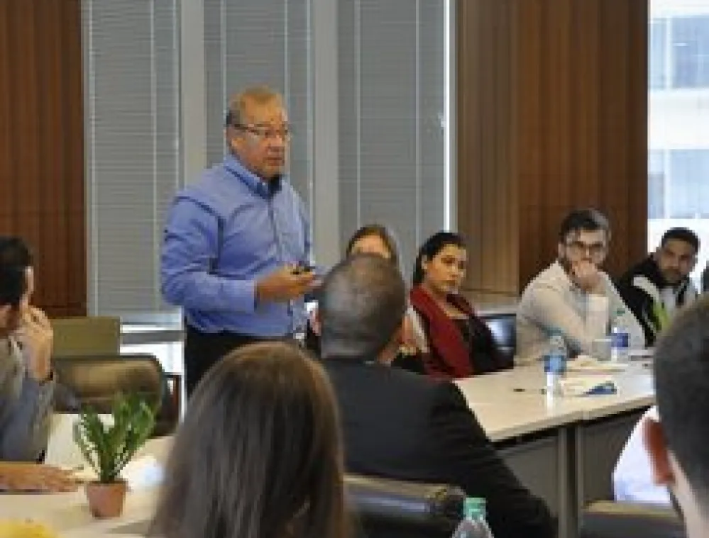 A man in a blue shirt stands and speaks to a group of people seated around a conference table in a modern office with large windows and blinds.