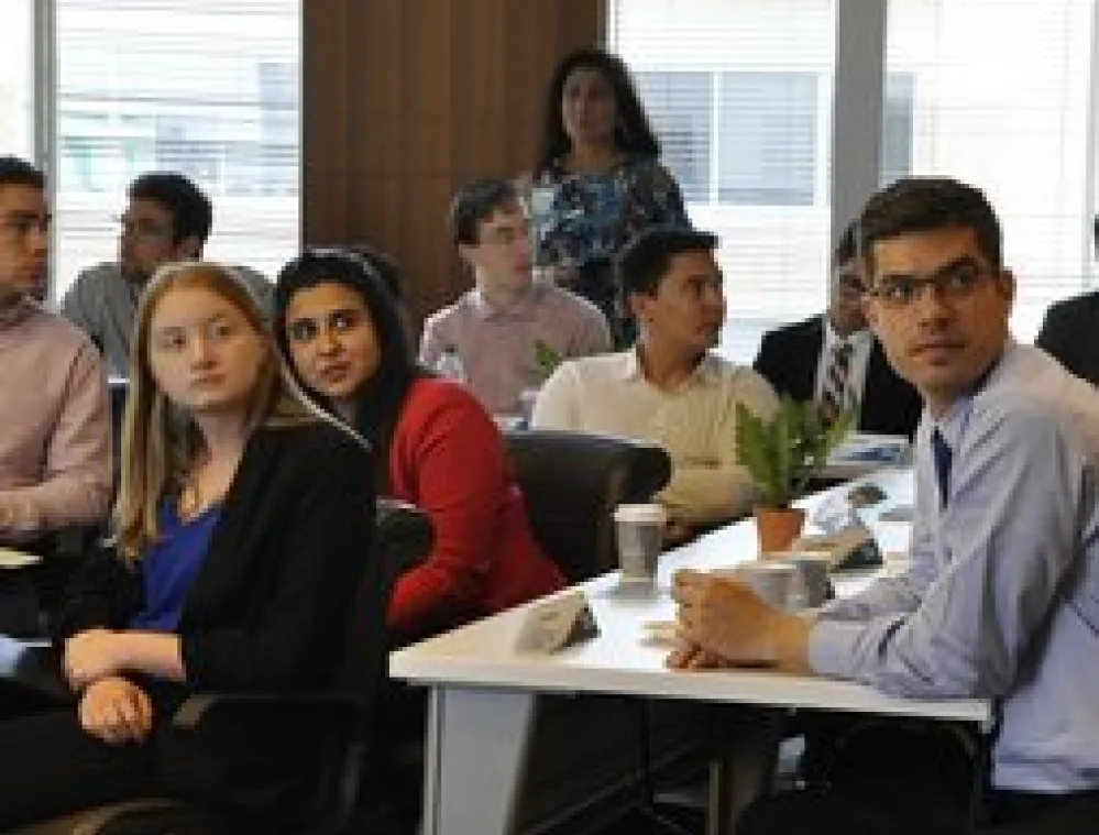 Group of diverse professionals seated at conference tables in a modern office, attentively listening to a presentation. Natural light comes through large windows, and nameplates, coffee cups, and a plant are visible on the tables.