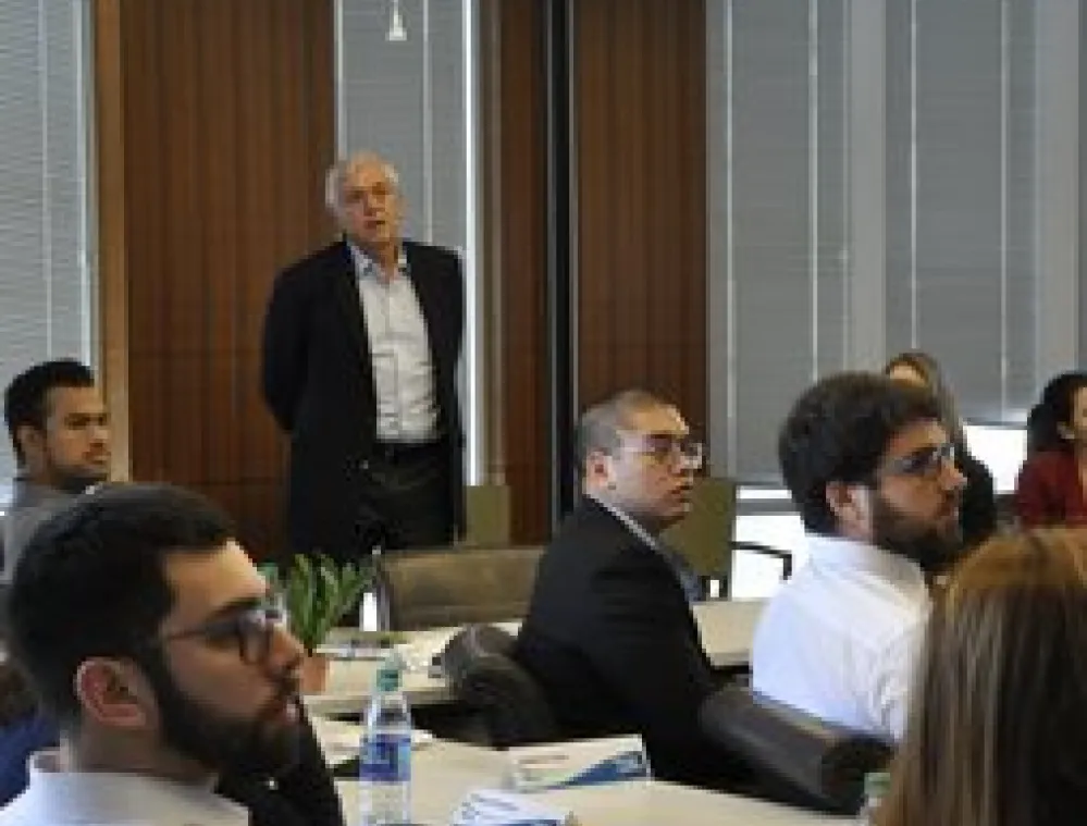 A group of people attentively listening to a speaker in a modern conference room with large windows and wooden panels.