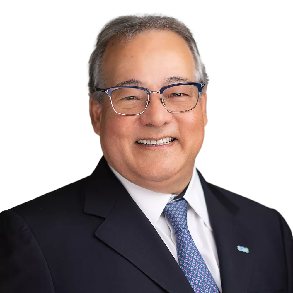 A man with short gray hair and glasses is smiling, wearing a dark suit, white shirt, and patterned tie.