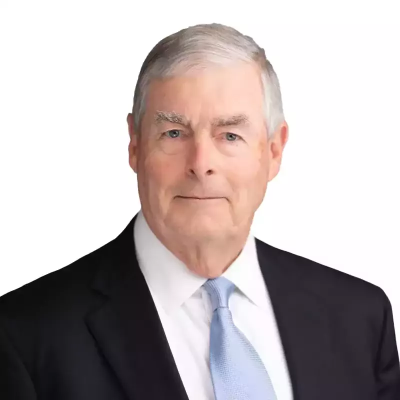 An older man with gray hair wearing a black suit, white shirt, and light blue tie, set against a plain white background.