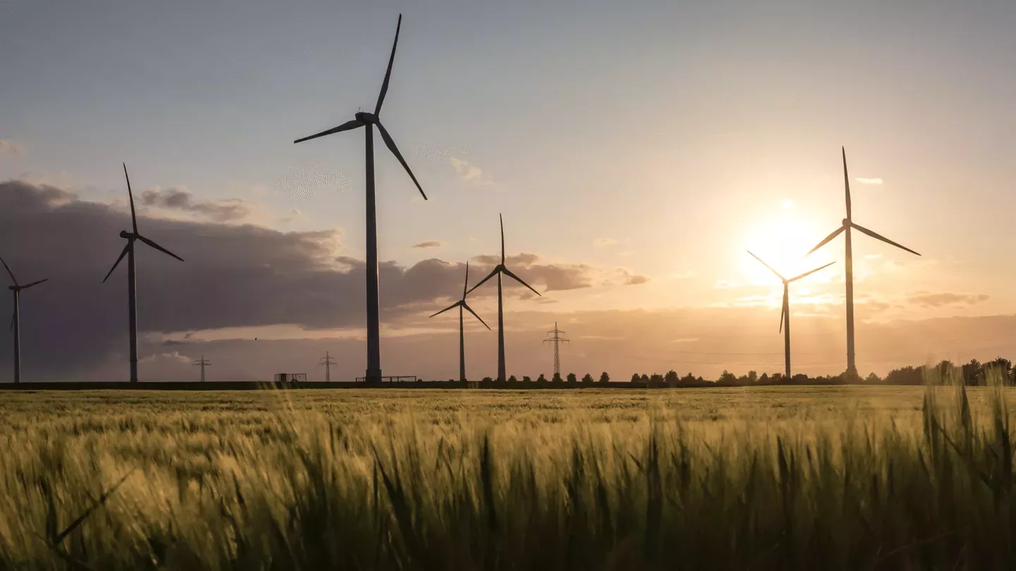 Wind turbines in a field during sunset, with a golden sky and scattered clouds. The sun is partially hidden behind one of the turbines, casting a warm glow over the landscape.