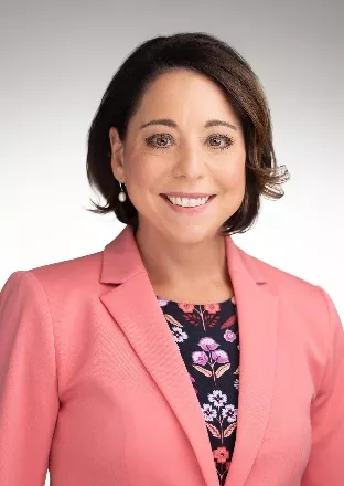 A woman with short dark hair smiles at the camera. She is wearing a coral blazer over a floral-patterned blouse. The background is a gradient of white to light gray.