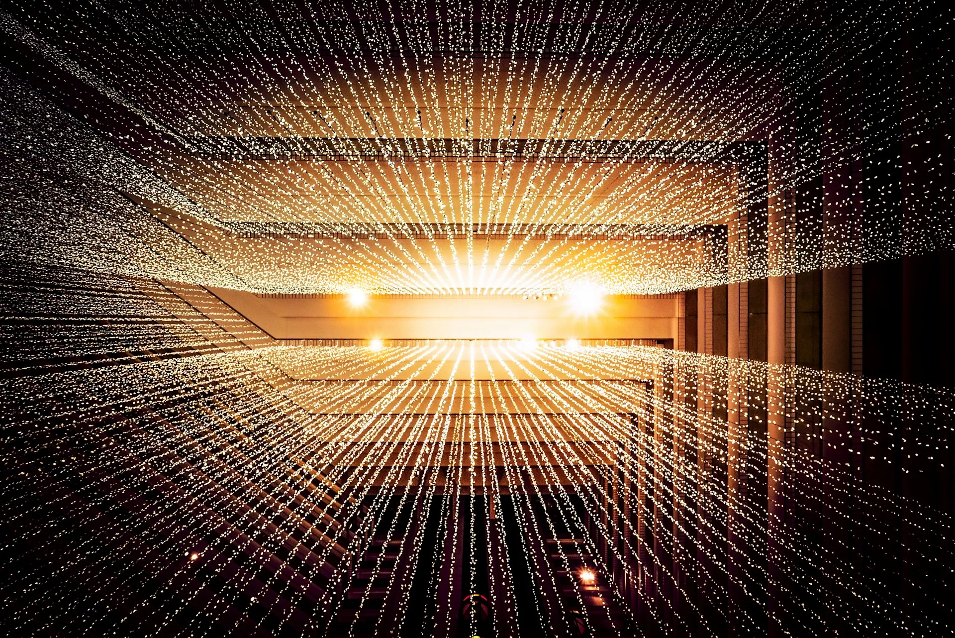 Rows of small white lights suspended in a geometric pattern inside a modern building, creating a radiant, starburst effect with warm lighting in the background.