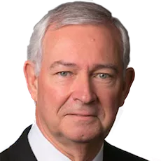 Portrait of an older man with short gray hair, wearing a dark suit and white shirt. He has a neutral expression and light-colored eyes.