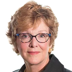 Portrait of a person with short curly hair, wearing glasses and a dark blazer, looking directly at the camera.